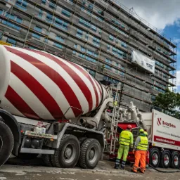 Ciężarówka z betonomieszarką w czerwono-białe pasy na tle budynku w rusztowaniach i dwóch pracowników budowlanych w odblaskowych ubraniach.