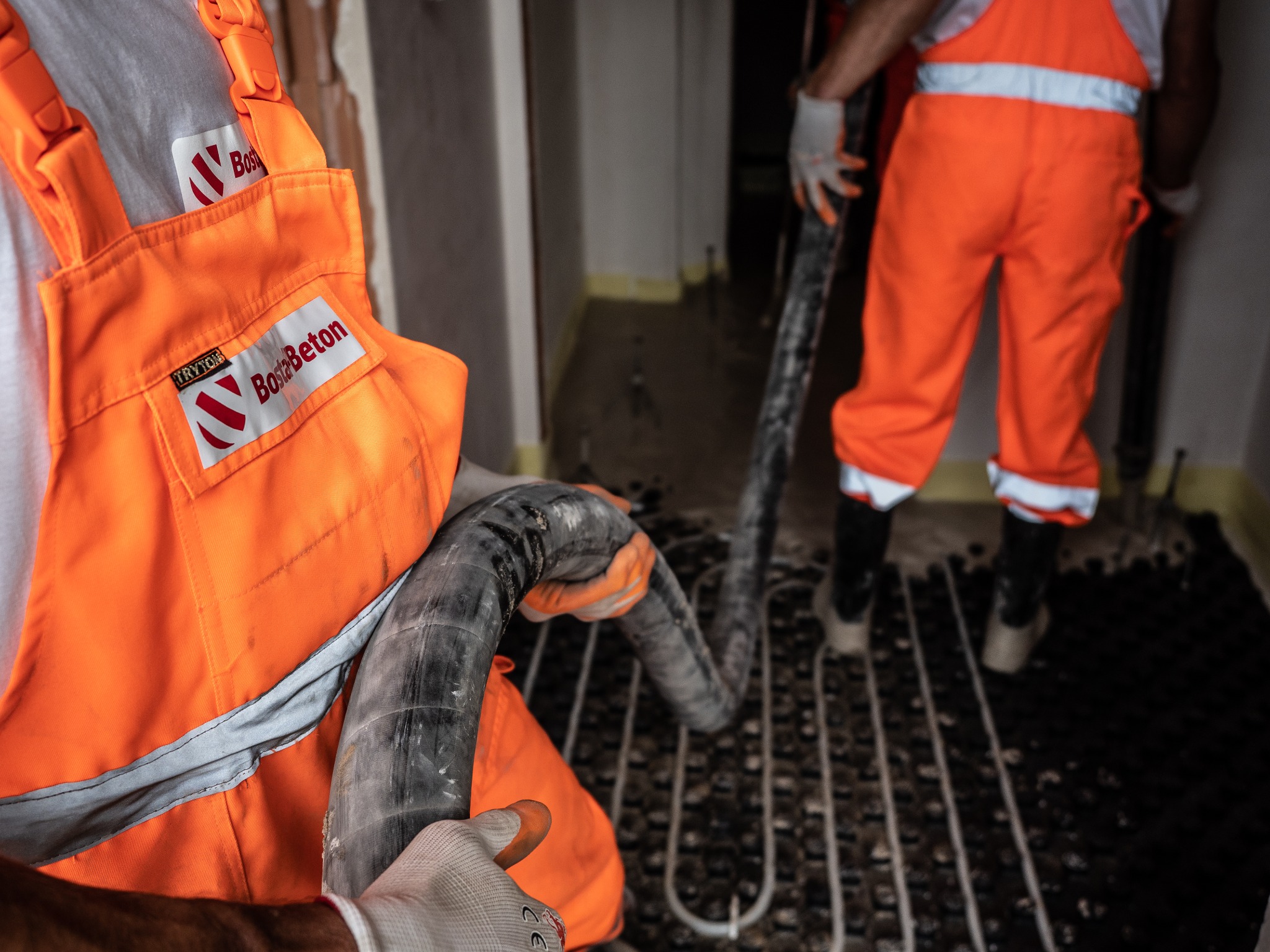 Pracownik w pomarańczowym kombinezonie z logo firmy 'Bosta Beton' w trakcie wylewania posadzki na ogrzewanie podłogowe przy użyciu węża. Widoczne fragmenty wylewki na czarnej macie.
