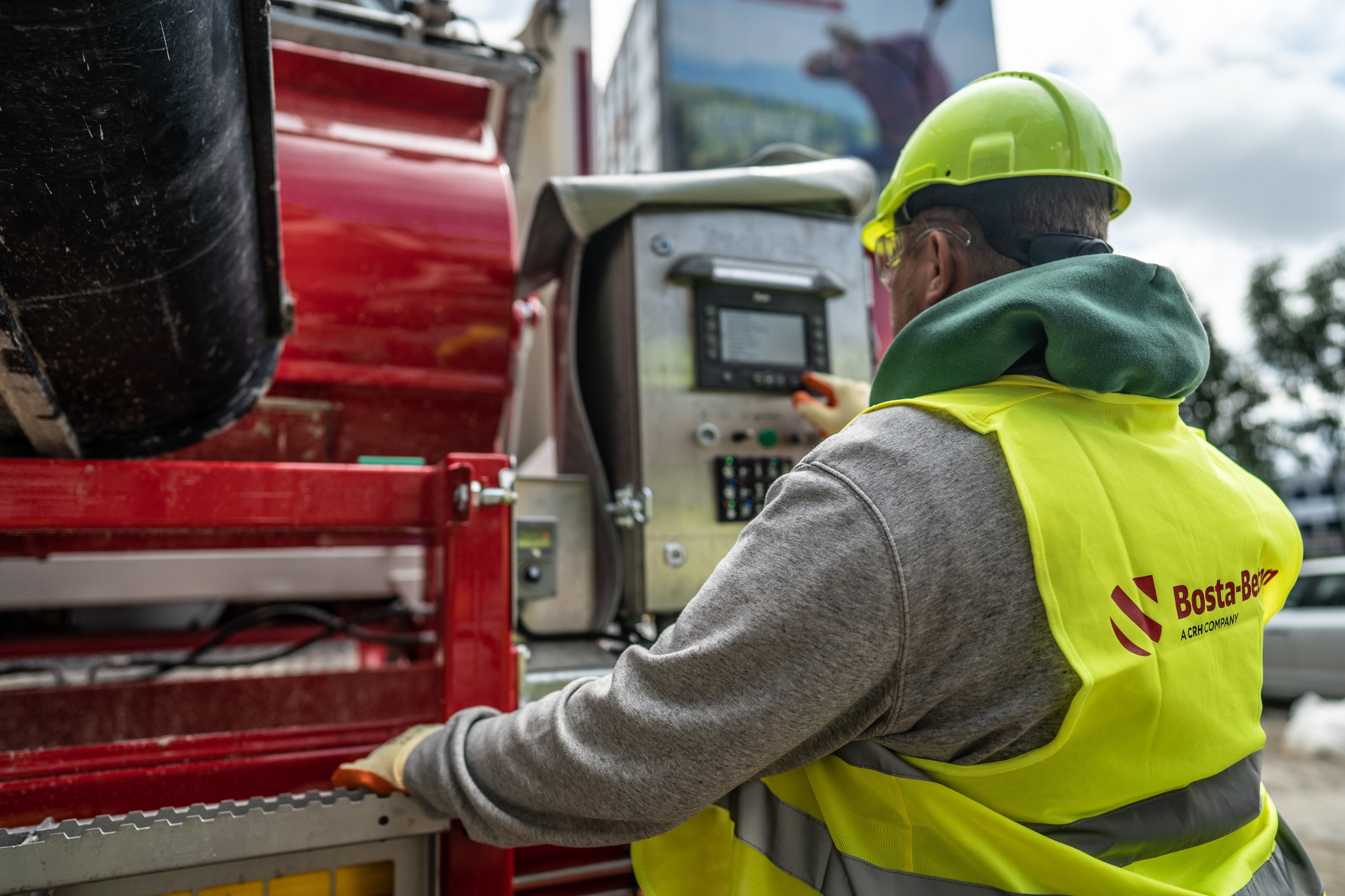 Pracownik w kasku i kamizelce odblaskowej obsługuje panel sterowania maszyny do wylewania posadzek, widoczny czerwony zbiornik i logo firmy Bosta-Bet.