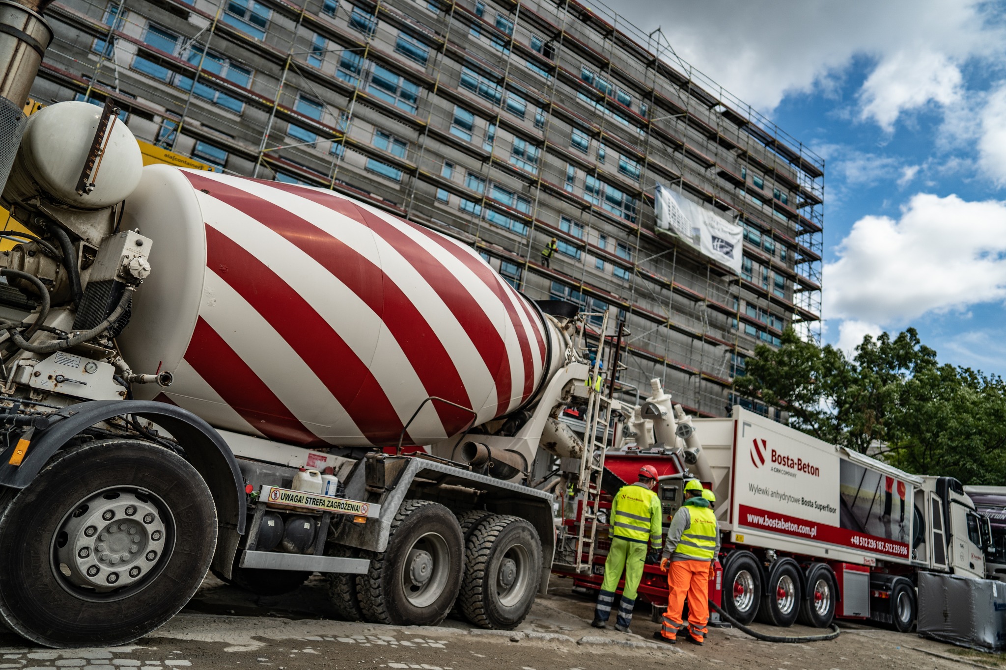 Ciężarówka z betonomieszarką w czerwono-białe pasy na tle budynku w rusztowaniach i dwóch pracowników budowlanych w odblaskowych ubraniach.