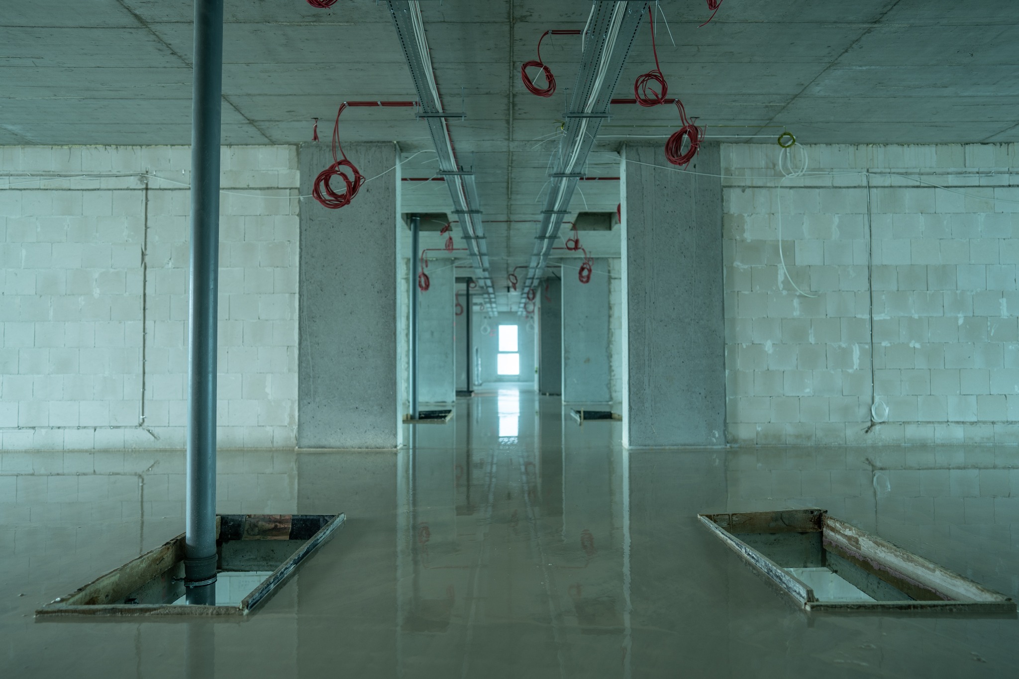 Wnętrze w trakcie wylewania posadzki, widoczne otwory w podłodze z wystającymi rurami, surowy stan ścian z bloczków i instalacje pod sufitem.