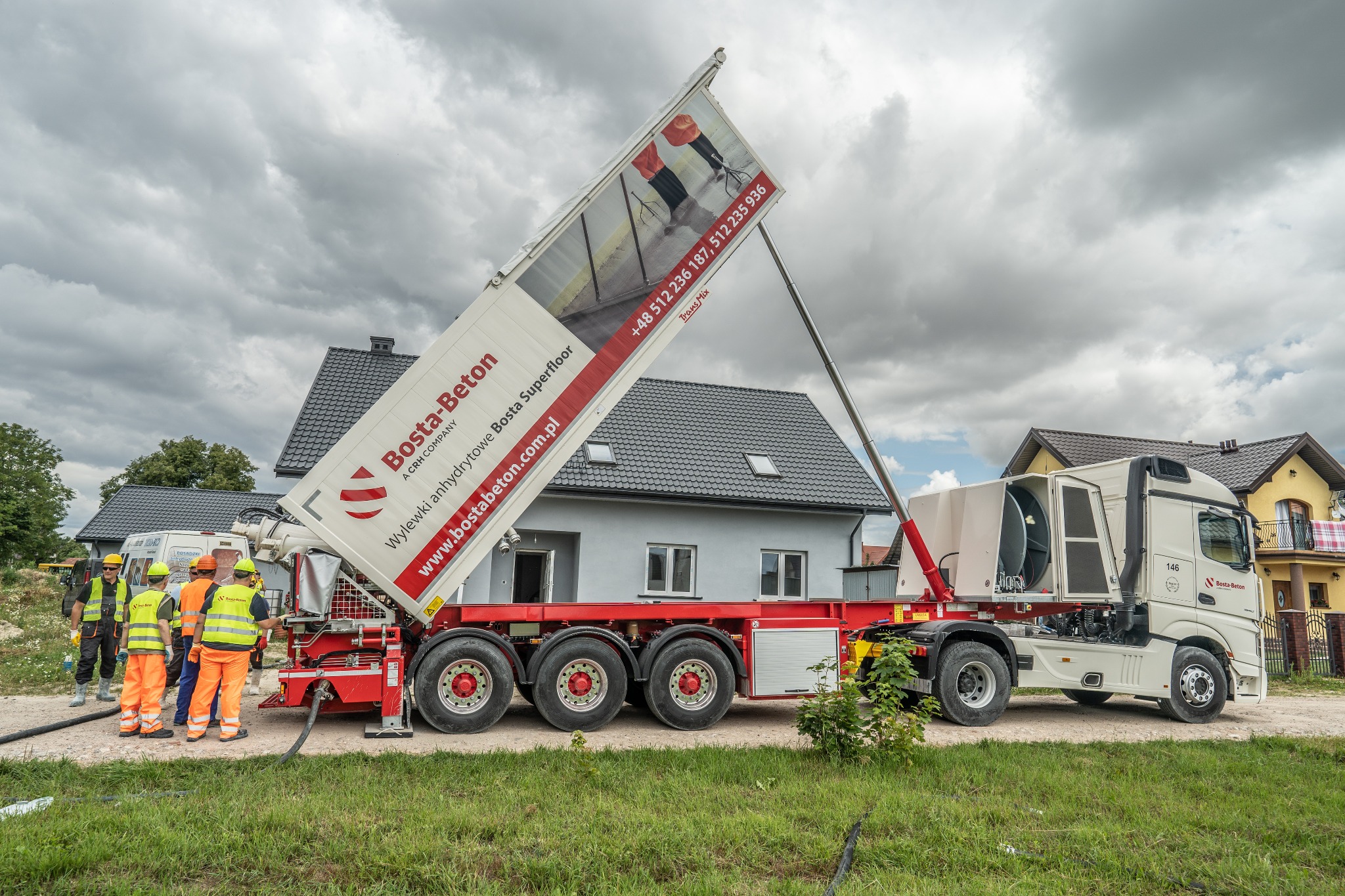 Ciężarówka z podnośnikiem z reklamą wylewek anhydrytowych Bosta-Beton na tle budynków mieszkalnych i grupy robotników w odblaskowych kamizelkach podłączających wąż.