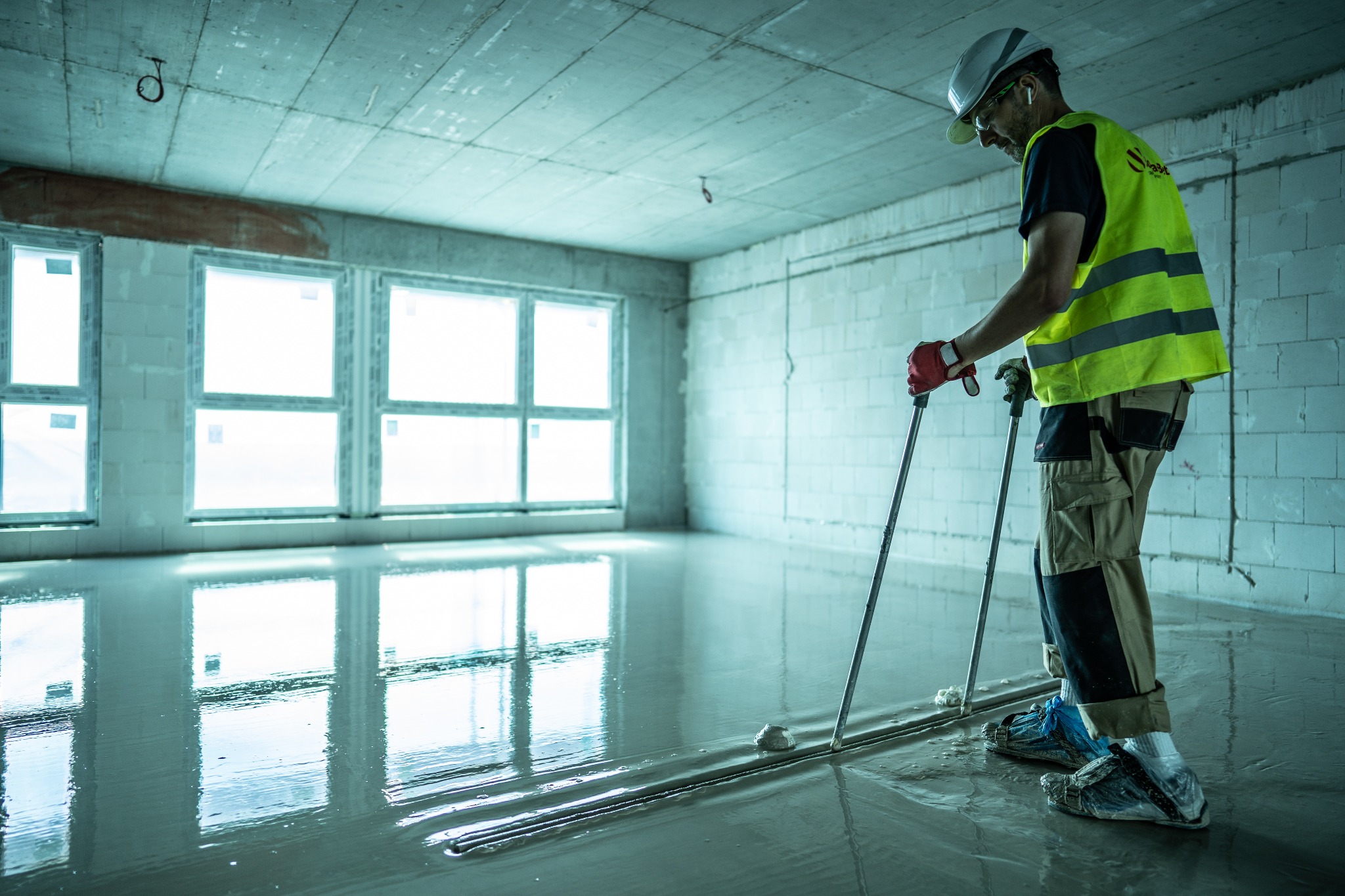Pracownik w kasku i kamizelce odblaskowej rozprowadza masę na podłodze w surowym wnętrzu z oknami, używając specjalnych narzędzi; widoczne refleksy światła na mokrej powierzchni.