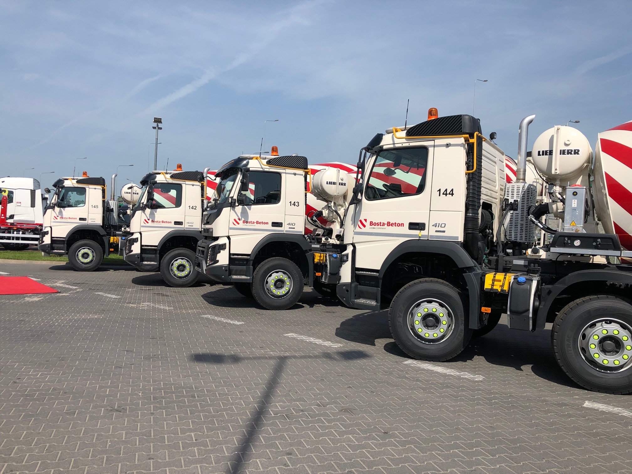 Flota białych betonomieszarek marki Liebherr z logo Bosta-Beton ustawiona w rzędzie na placu z kostki brukowej, widok z boku, błękitne niebo w tle.