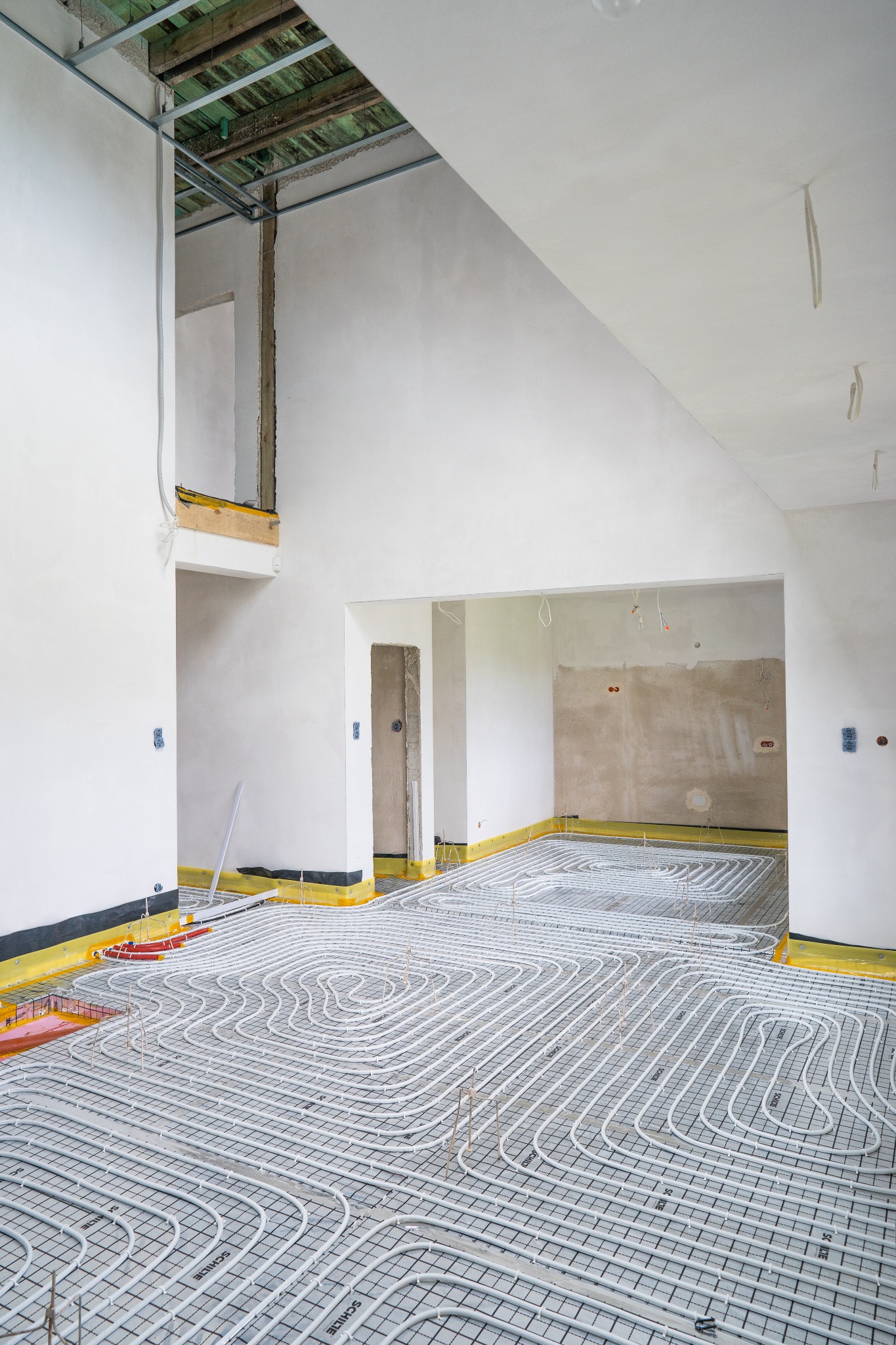 Wnętrze w trakcie montażu ogrzewania podłogowego: białe rury grzewcze ułożone na czarnej siatce, żółta folia izolacyjna przy ścianach, surowy stan ścian i sufitu z widocznymi elementami...