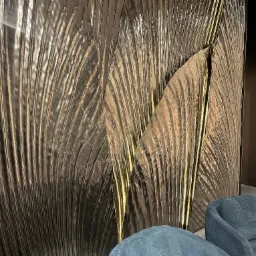 Dekoracyjna ściana z metalicznym efektem przypominającym liście, podświetlona złotymi akcentami, uzupełniona przez dwa turkusowe fotele.