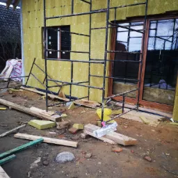 Budynek w trakcie ocieplania, widoczne rusztowanie, okna z ramą w kolorze brązowym, ściany pokryte żółtą wełną mineralną z widocznymi kołkami mocującymi, na ziemi porozrzucane materiały budowlane...