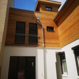 Nowoczesny budynek z elewacją z jasnego drewna, czarnymi oknami z balkonem, widok z dołu na błękitne niebo.