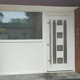 Białe drzwi wejściowe z przeszklonym panelem bocznym w nowoczesnym domu z cegły, widoczne odbicie fotografa w szybie.