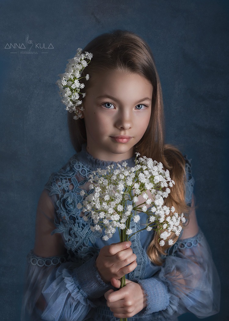 Portret dziewczynki w niebieskiej sukience z koronką, trzymającej bukiet białych kwiatów na tle teksturowanej ściany w odcieniach błękitu, z kwiatami we włosach i subtelnym oświetleniem.