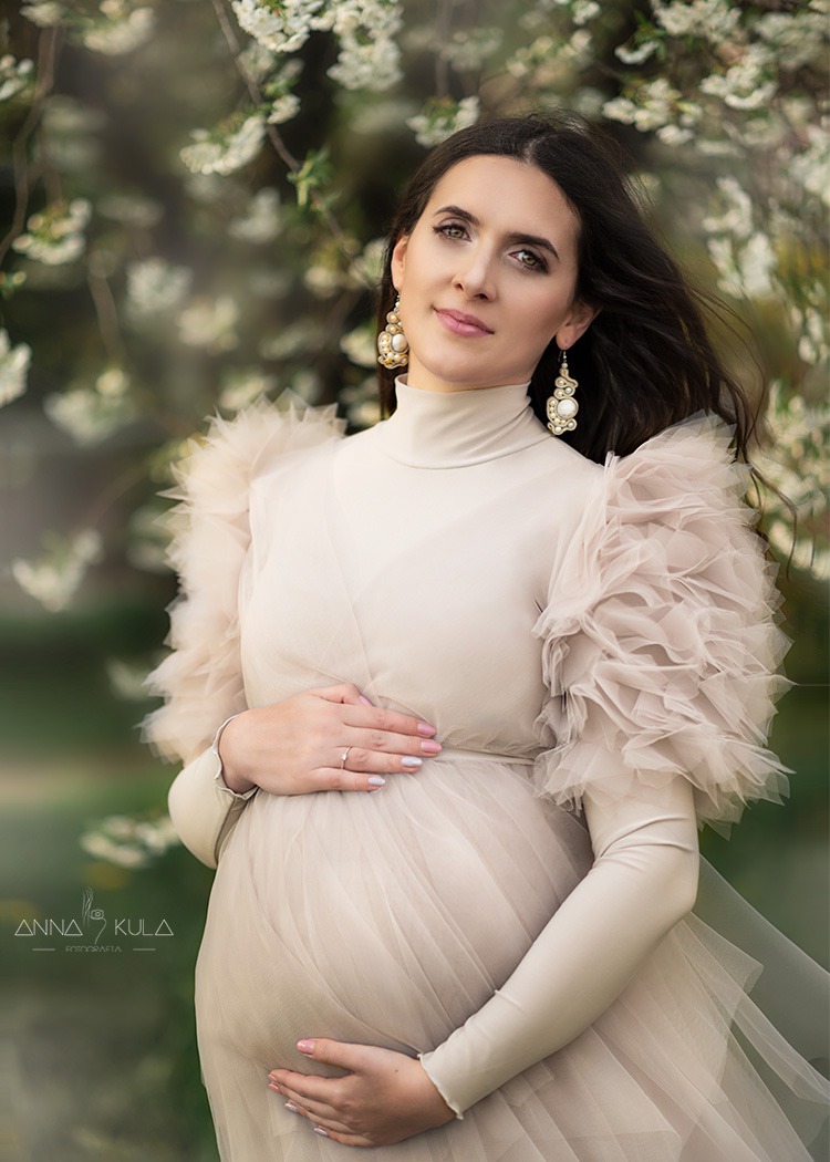 Kobieta w ciąży w beżowej sukni z falbanami na tle kwitnących gałęzi, delikatny uśmiech, duże kolczyki, dłonie na brzuchu.