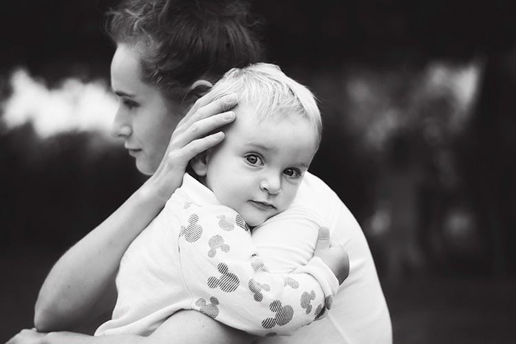 Czarno-białe zdjęcie matki obejmującej małe dziecko w ubraniu z motywem Myszki Miki, dziecko patrzy w obiektyw z delikatnym uśmiechem.