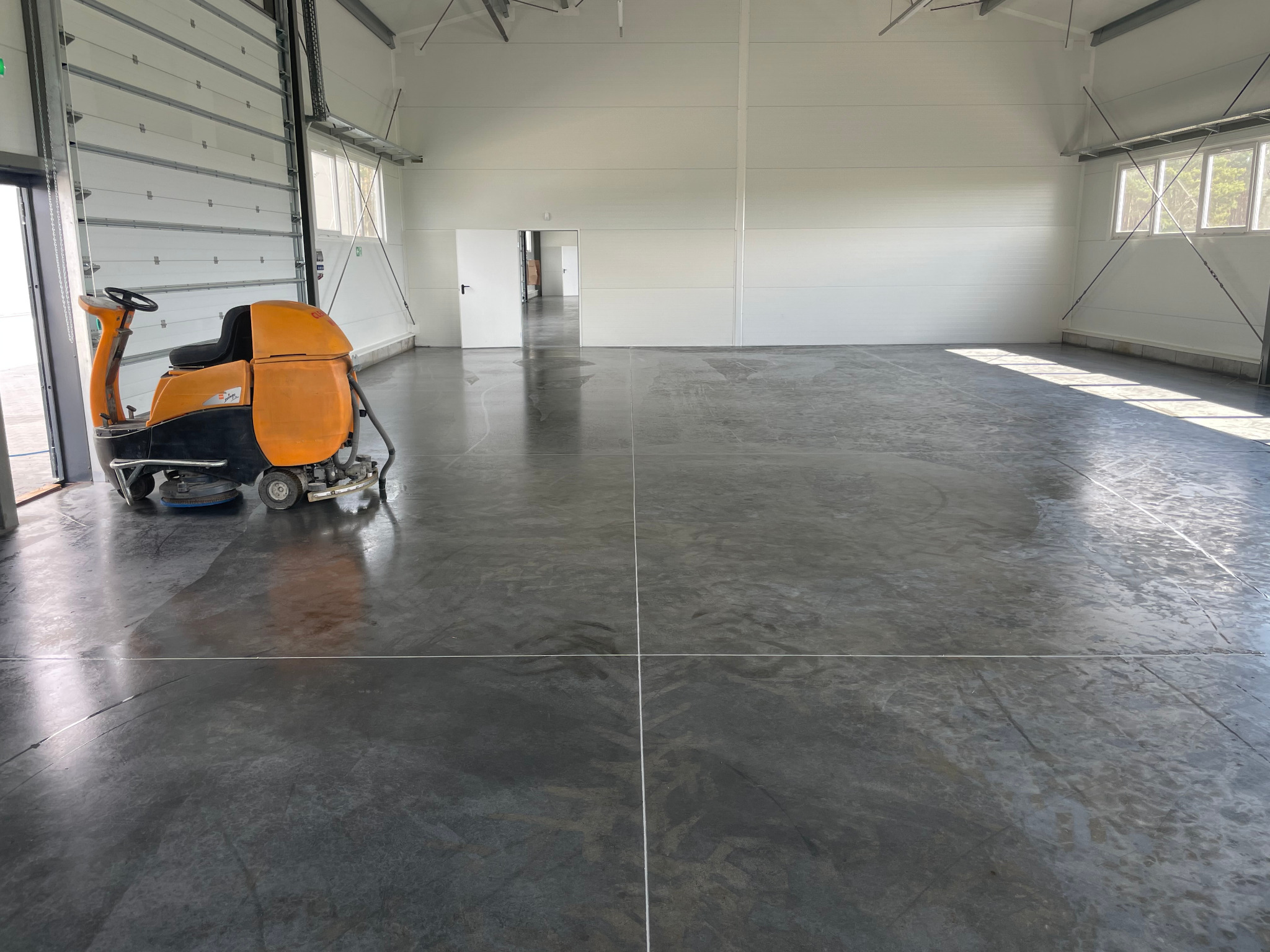 Pomarańczowa maszyna do mycia posadzek przemysłowych w hali z betonową podłogą i białymi ścianami, widoczne ślady po myciu.