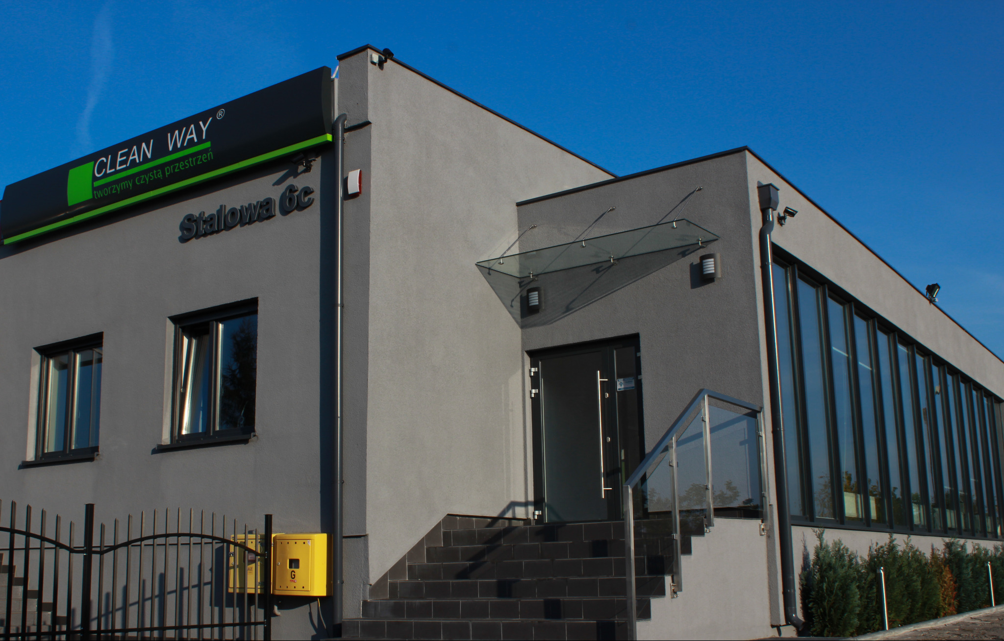 Szary budynek firmy Clean Way z dużymi oknami, wejściem ze schodami i szklanym daszkiem, widoczny szyld z nazwą firmy.