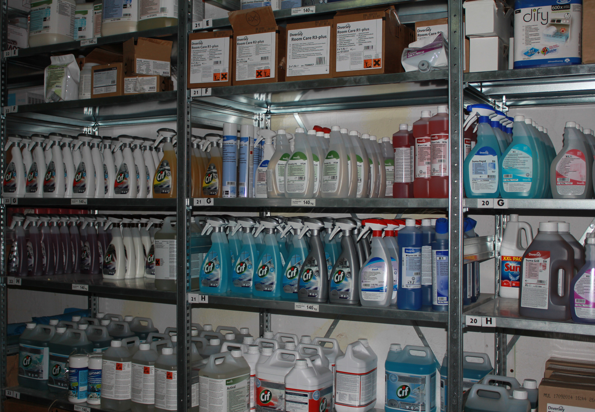 Półki magazynowe wypełnione różnorodnymi środkami czystości w butelkach i kanistrach, głównie marki Cif i Diversey, ułożone w rzędach na metalowych regałach.