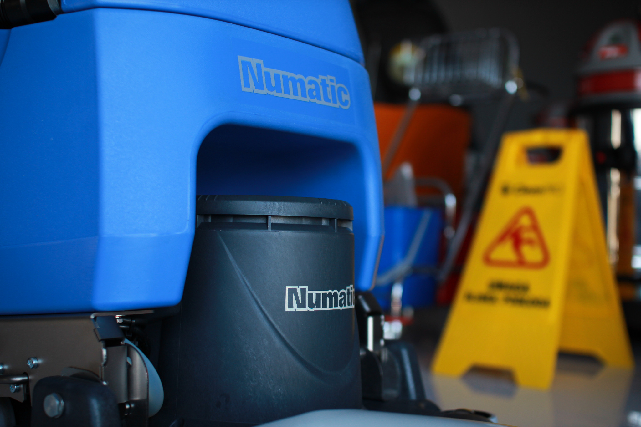 Niebieska maszyna czyszcząca Numatic z żółtym znakiem ostrzegawczym 'Uwaga śliska podłoga' w tle.