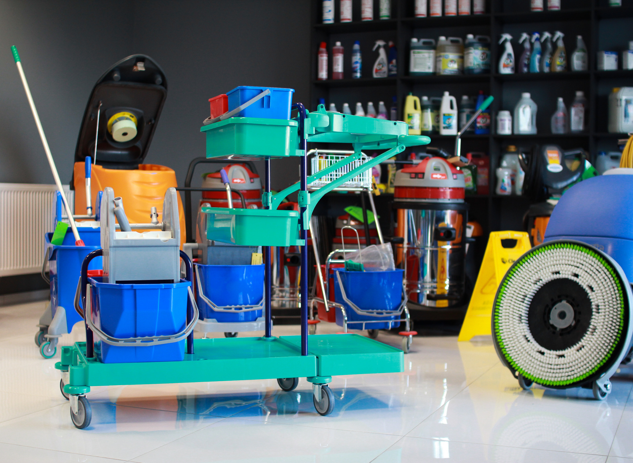 Wózki i maszyny do sprzątania z mopem i wiadrami na błyszczącej podłodze, półki z detergentami w tle.
