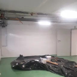 Przygotowania do malowania pomieszczenia: rozłożona folia ochronna, puszki z farbą, wałek malarski i kartonowa tuba na podłodze, widoczne rury pod sufitem i wieszak.