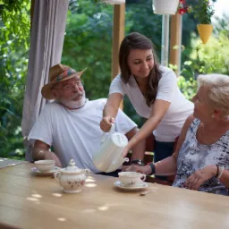 Młoda kobieta nalewa napój z białego dzbanka do filiżanki siedzącej przy stole starszej pani, obok siedzi starszy pan w kapeluszu na tle zieleni.