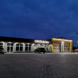 Budynek stacji kontroli pojazdów i serwisu OSKP Anika o zmierzchu, z zaparkowanymi samochodami na brukowanym placu.