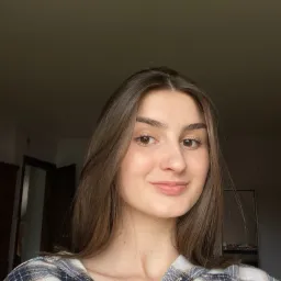 Portret uśmiechniętej brunetki w kraciastej koszuli na tle ciemnego wnętrza.