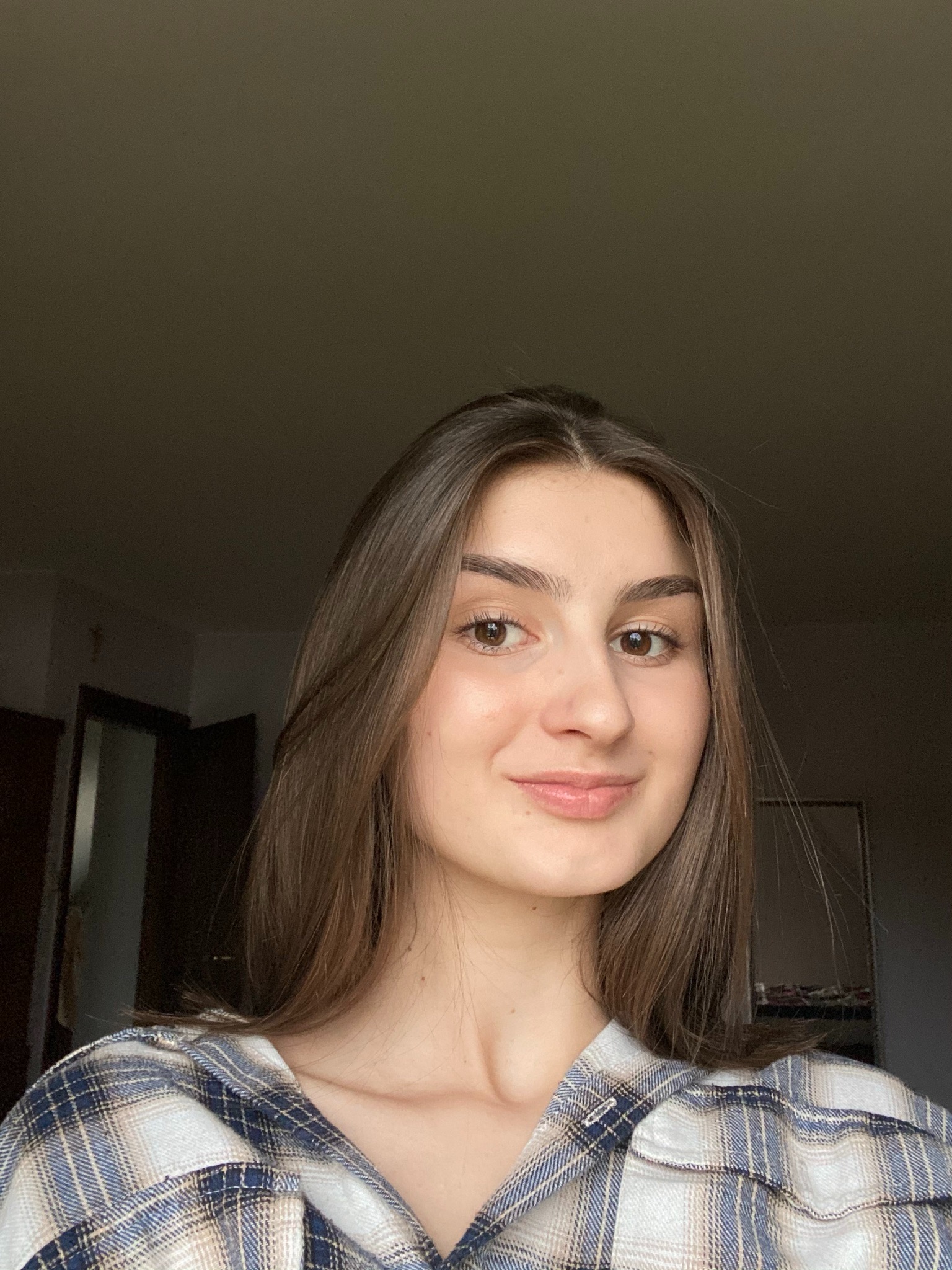 Portret uśmiechniętej brunetki w kraciastej koszuli na tle ciemnego wnętrza.