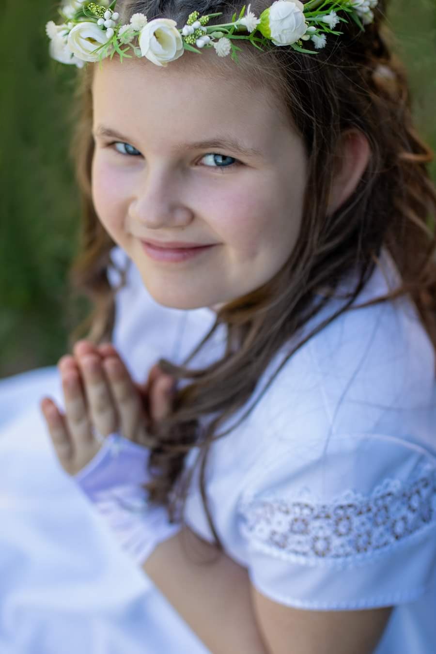 Dziewczynka w białej sukience i wianku z białych kwiatów, z dłońmi złożonymi do modlitwy, delikatny uśmiech, błękitne oczy, zielone tło.