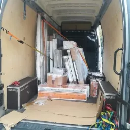 Wnętrze vana załadowane oknami i drzwiami zabezpieczonymi pasami transportowymi, widoczne kartony i skrzynie z logo Knauftec.