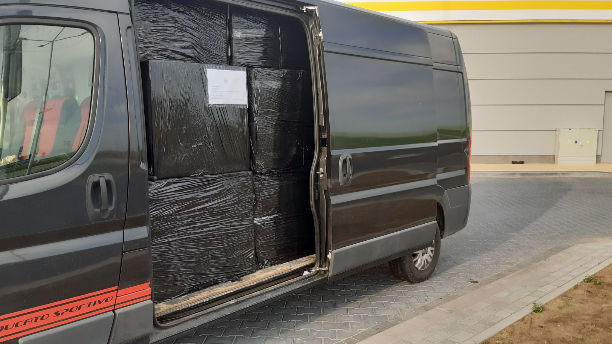 Czarny bus Ducato Sportivo z otwartymi drzwiami bocznymi, wypełniony po brzegi czarnymi, szczelnie zapakowanymi paczkami, widoczna naklejka adresowa na jednej z paczek.