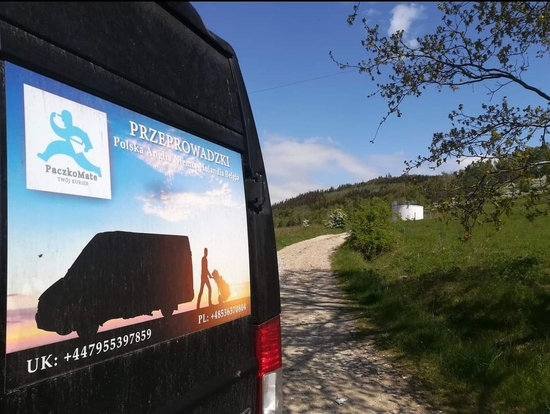 Reklama firmy przeprowadzkowej na boku czarnego busa, z numerami kontaktowymi dla Wielkiej Brytanii i Polski, na tle wiejskiej drogi i wzgórz.