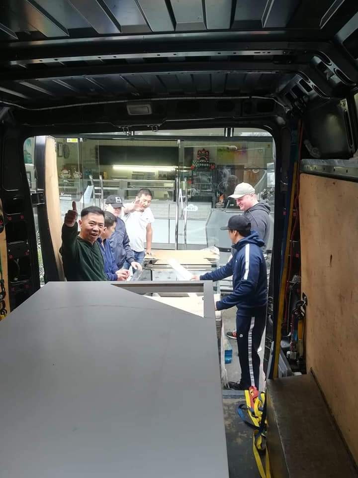 Wnętrze busa z grupą osób ładujących duży, szary element konstrukcyjny; jeden mężczyzna unosi kciuk w górę, wyrażając aprobatę.