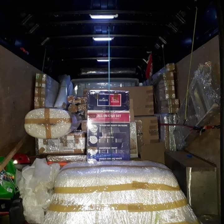 Wnętrze busa wypełnione po brzegi zapakowanymi przedmiotami, w tym pudełko z zestawem ceramiki sanitarnej, owinięte folią meble i kartony, zabezpieczone taśmą, oświetlenie sufitowe w tle.