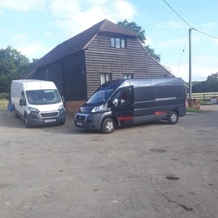 Dwa busy transportowe, jeden biały i jeden czarny z otwartymi drzwiami, zaparkowane przed drewnianym budynkiem gospodarczym w słoneczny dzień.