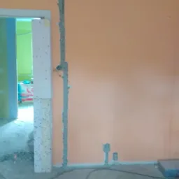 Ściana w trakcie remontu z wyżłobionymi bruzdami pod przewody elektryczne, widoczne kable i gruz na podłodze, pomarańczowa ściana, niedokończona futryna drzwi.