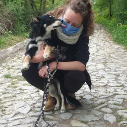 Kobieta w masce przykuca na brukowanej ścieżce w parku, trzymając czarno-białego psa, który liże jej twarz.