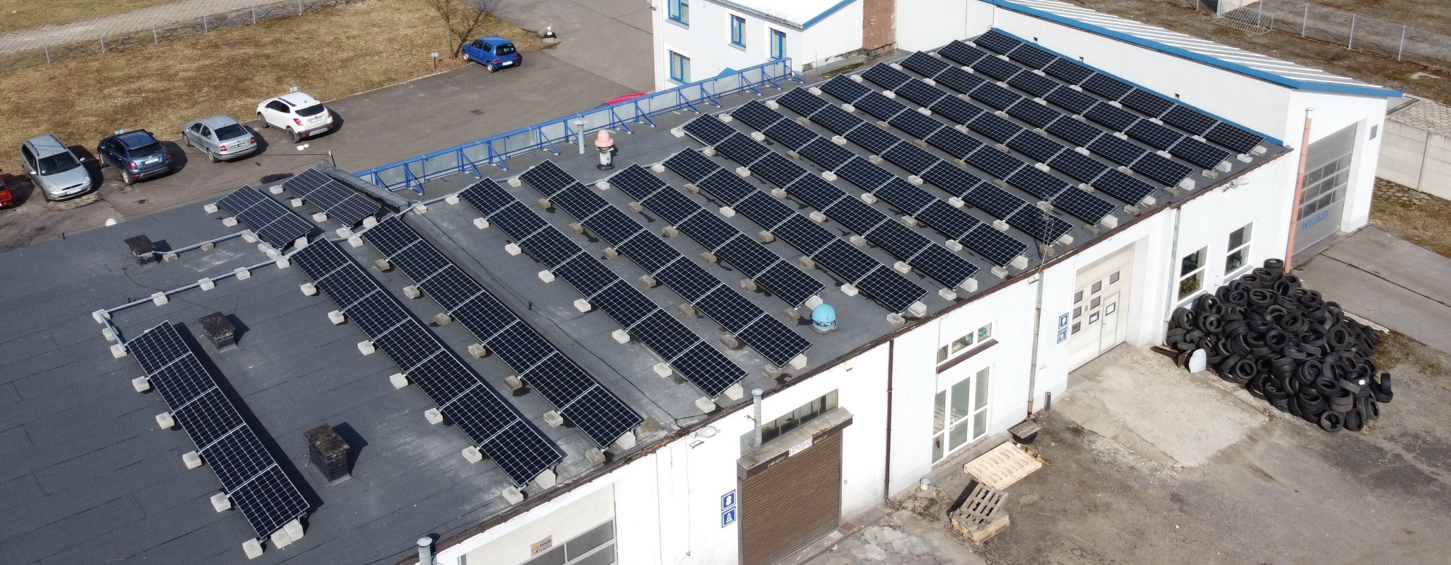 Widok z góry na budynek z zainstalowanymi panelami słonecznymi na dachu, obok sterta opon i zaparkowane samochody.