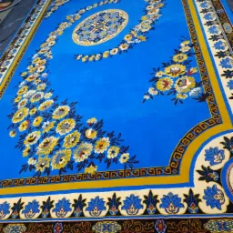 Duży, prostokątny dywan w odcieniach błękitu z bogatym, kwiatowym wzorem i beżowymi frędzlami, leżący na czarnej folii ochronnej.