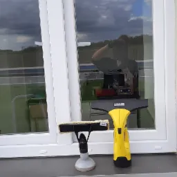 Ż&oacute;łty elektryczny odkurzacz do okien K&auml;rcher i spryskiwacz z mikrofibrą stoją na parapecie okna z białymi ramami, odbijającymi niebo i otoczenie.