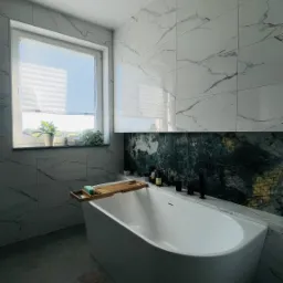 Nowoczesna łazienka z wolnostojącą wanną, marmurowe płytki ścienne, drewniana półka na wannie z mydłem, roślina w doniczce na parapecie, szary dywanik podłogowy, czarne baterie i oświetlenie.