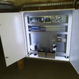 Otwarta szafa sterownicza z modułami elektronicznymi, zasilaczem i akumulatorami, zamontowana w pomieszczeniu z widocznymi instalacjami pod sufitem.