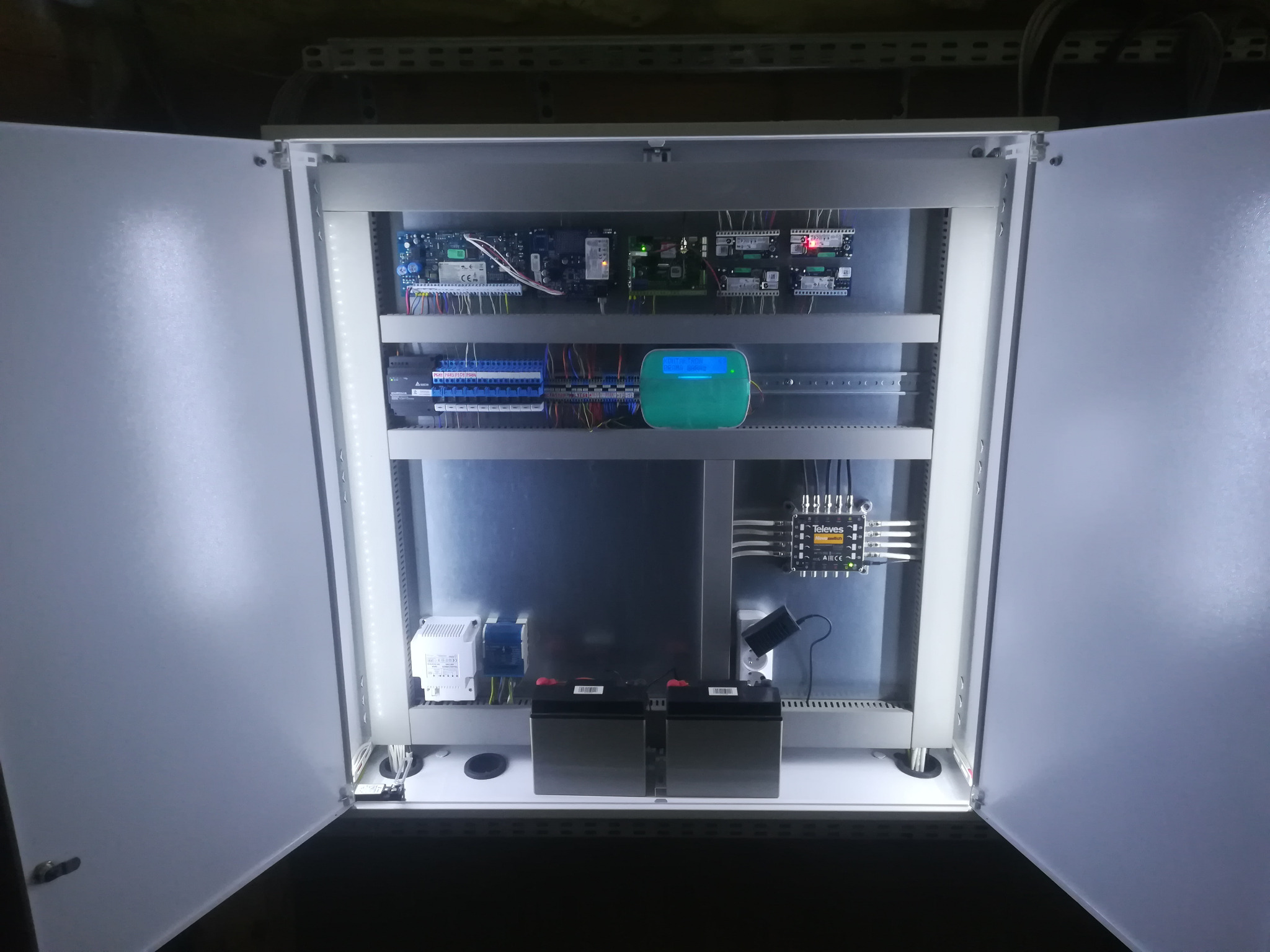 Otwarta szafa sterownicza z widocznymi modułami elektronicznymi, przewodami, akumulatorami i elementami zasilania, oświetlona wewnętrznym światłem LED.