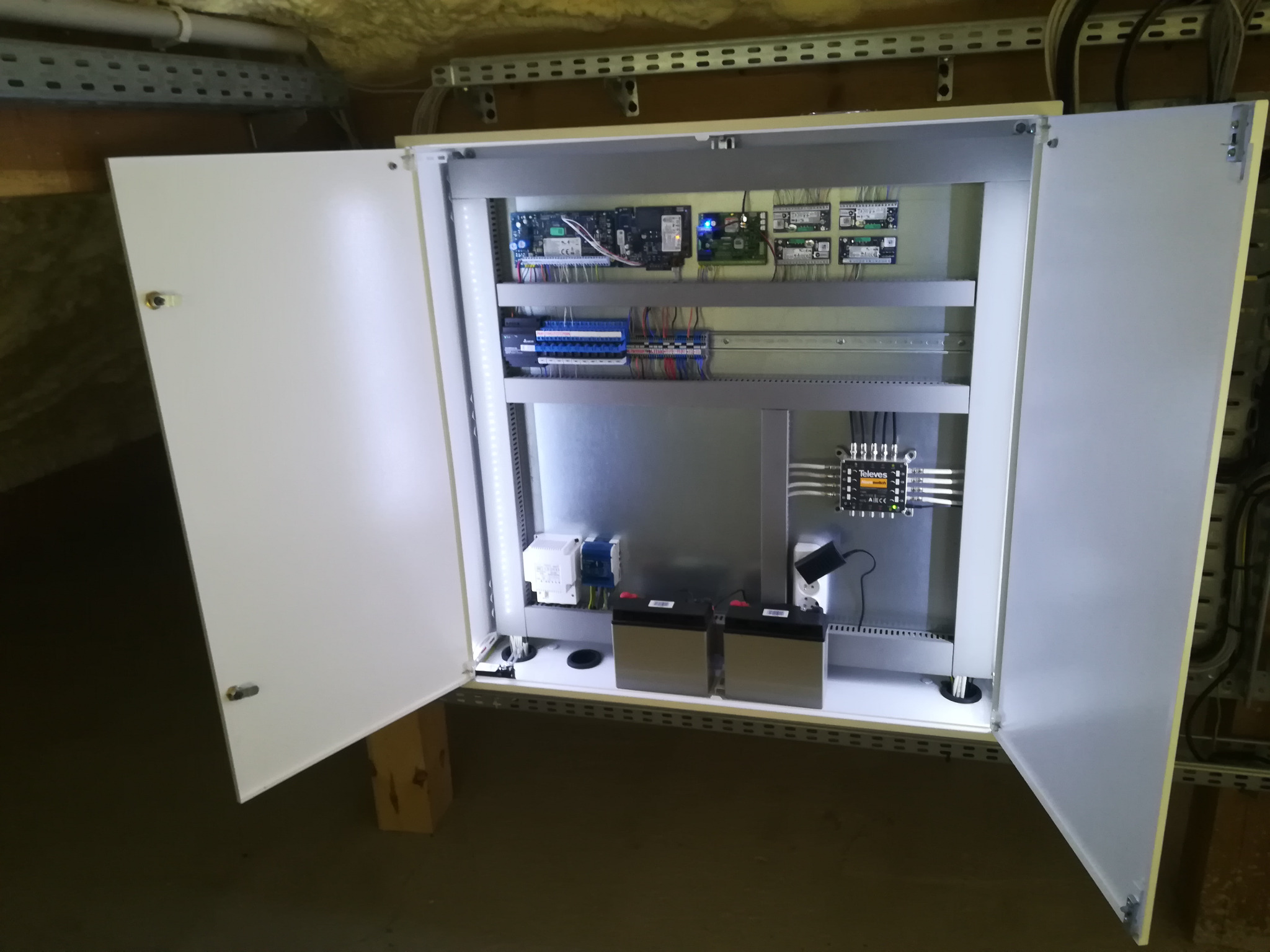 Otwarta szafa sterownicza z modułami elektronicznymi, zasilaczem i akumulatorami, zamontowana w pomieszczeniu z widocznymi instalacjami pod sufitem.