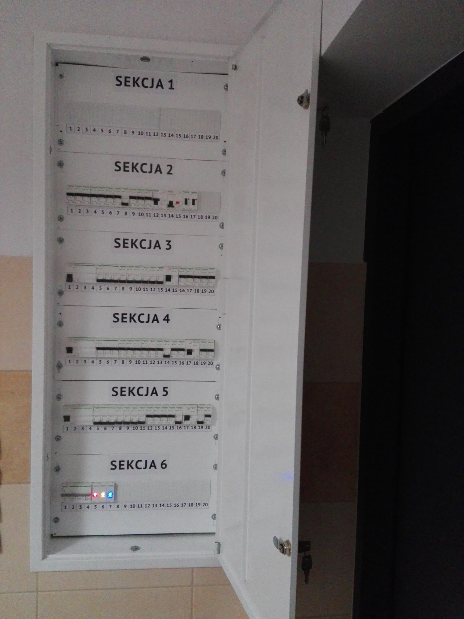 Otwarta, biała skrzynka rozdzielcza z opisem sekcji i numeracją, widoczne wyłączniki, diody sygnalizacyjne w sekcji 6, kluczyk w zamku.