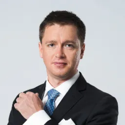 Portret mężczyzny w czarnym garniturze, białej koszuli z mankietami i niebieskim krawacie w kratę, z chusteczką w kieszonce, na jasnoszarym tle.