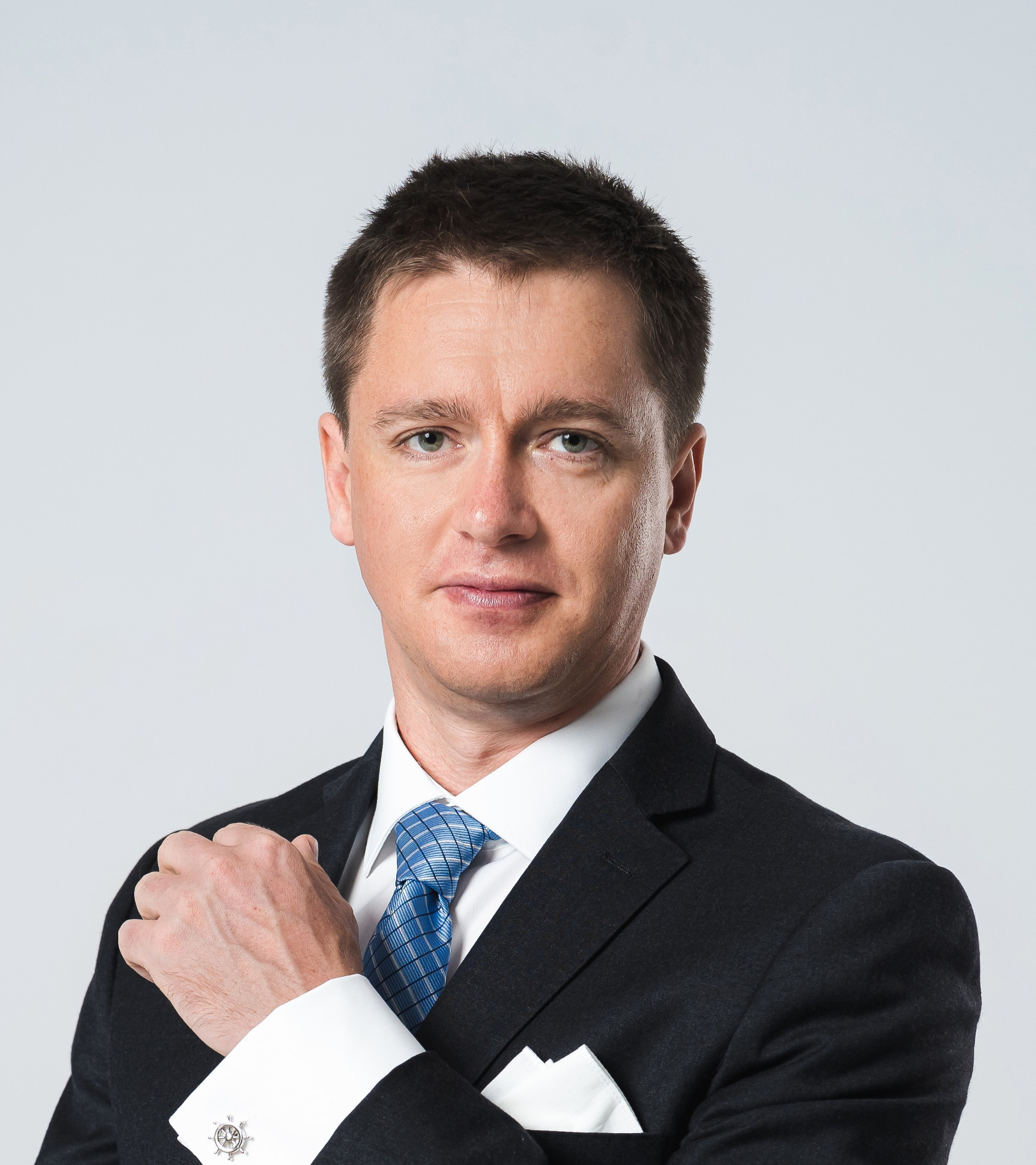 Portret mężczyzny w czarnym garniturze, białej koszuli z mankietami i niebieskim krawacie w kratę, z chusteczką w kieszonce, na jasnoszarym tle.