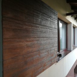 Nabogato Marcin Górecki - Elewacja domu z tynkiem drewnopodobnym, imitującym poziome deski w odcieniach brązu, sąsiadująca z oknami i fragmentem ściany z cegły.