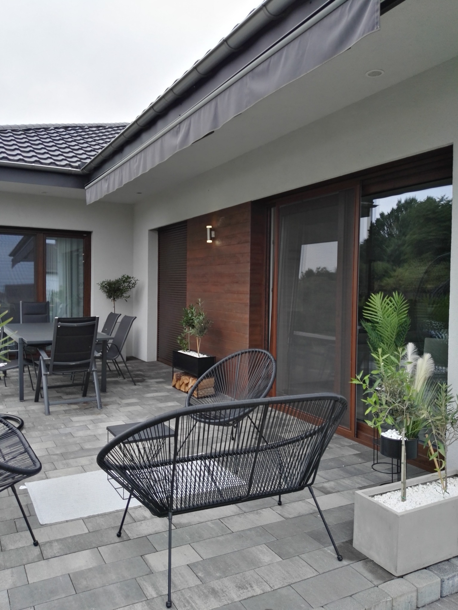 Nowoczesny taras z tynkiem drewnopodobnym na elewacji, meble ogrodowe z czarnego rattanu, szara kostka brukowa i rośliny w donicach.
