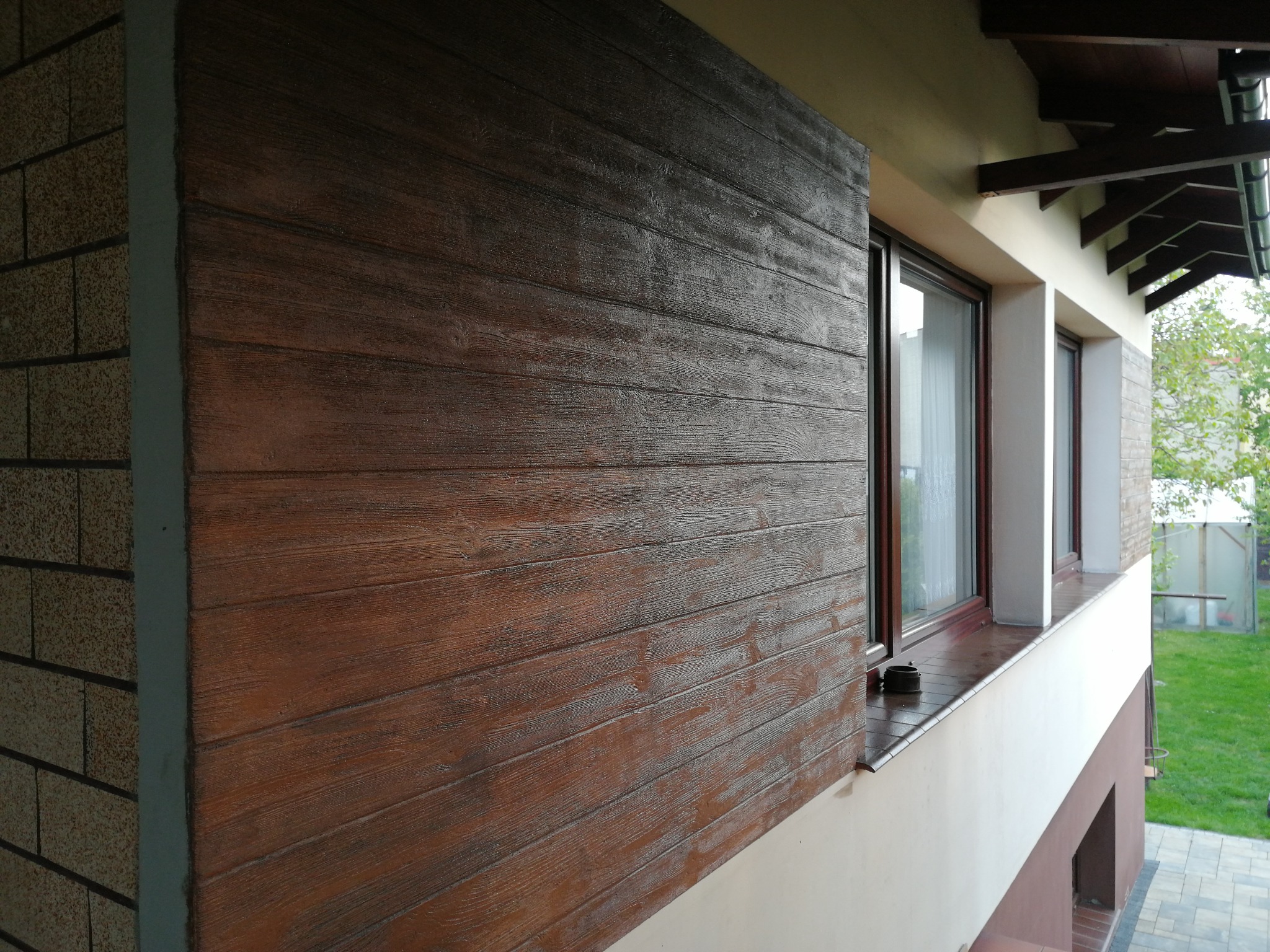 Elewacja domu z tynkiem drewnopodobnym, imitującym poziome deski w odcieniach brązu, sąsiadująca z oknami i fragmentem ściany z cegły.