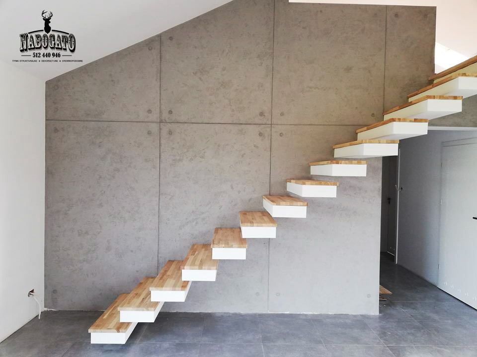 Nowoczesne schody dywanowe z drewnianymi stopniami i białymi podstopnicami, zamontowane przy ścianie z betonu architektonicznego w minimalistycznym wnętrzu.