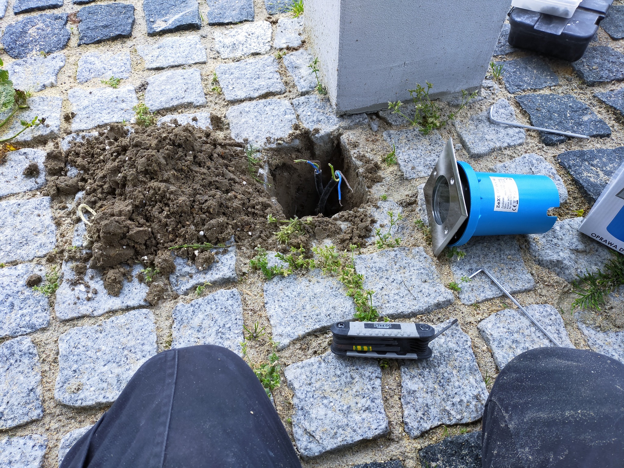 Przygotowanie do montażu lampy ogrodowej: wykopany otwór w kostce brukowej z widocznymi przewodami, obok leży niebieska obudowa lampy, narzędzia i odkopana ziemia.